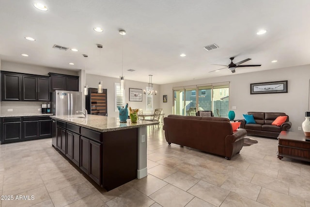 kitchen with light stone counters, hanging light fixtures, stainless steel fridge, an island with sink, and decorative backsplash