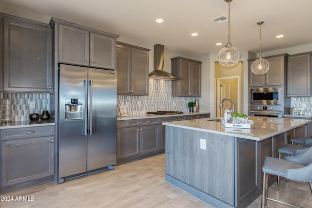 kitchen with appliances with stainless steel finishes, tasteful backsplash, wall chimney exhaust hood, decorative light fixtures, and an island with sink