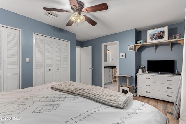 bedroom featuring light wood finished floors, visible vents, a ceiling fan, ensuite bathroom, and multiple closets