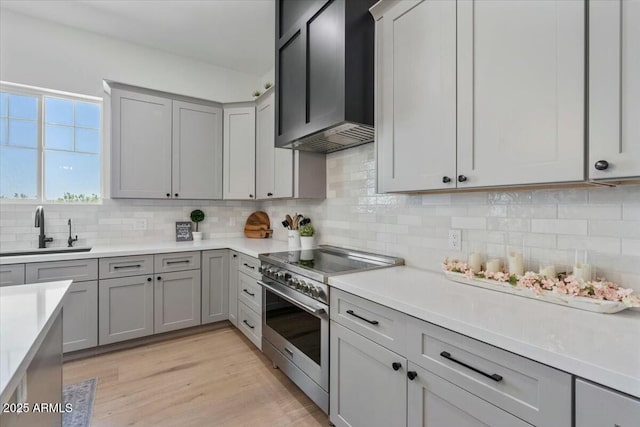 kitchen featuring stainless steel range, a sink, wall chimney exhaust hood, gray cabinetry, and tasteful backsplash