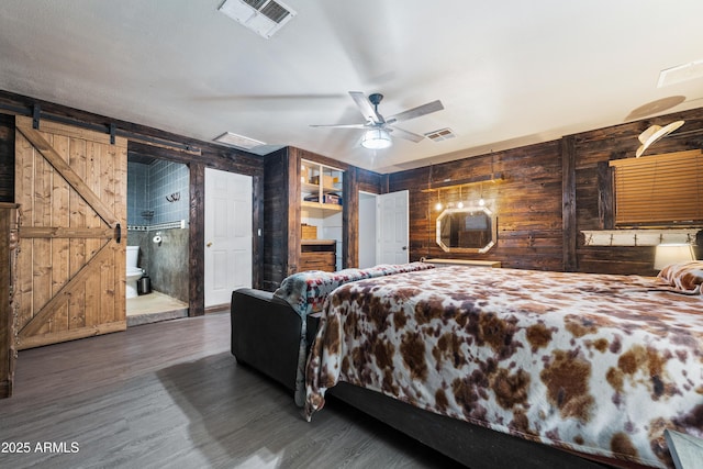 bedroom with ceiling fan, dark hardwood / wood-style flooring, a barn door, ensuite bath, and wooden walls