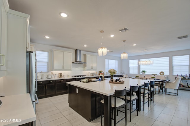 kitchen featuring decorative light fixtures, stainless steel fridge, white cabinets, a kitchen island with sink, and wall chimney exhaust hood