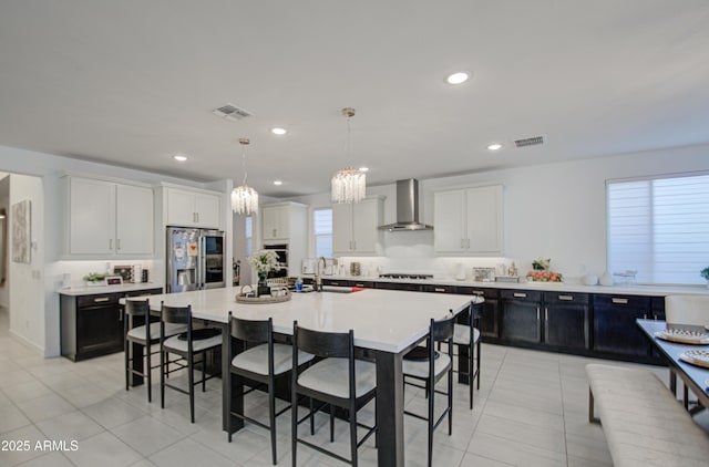 kitchen featuring hanging light fixtures, a kitchen island with sink, stainless steel fridge with ice dispenser, gas stovetop, and wall chimney range hood