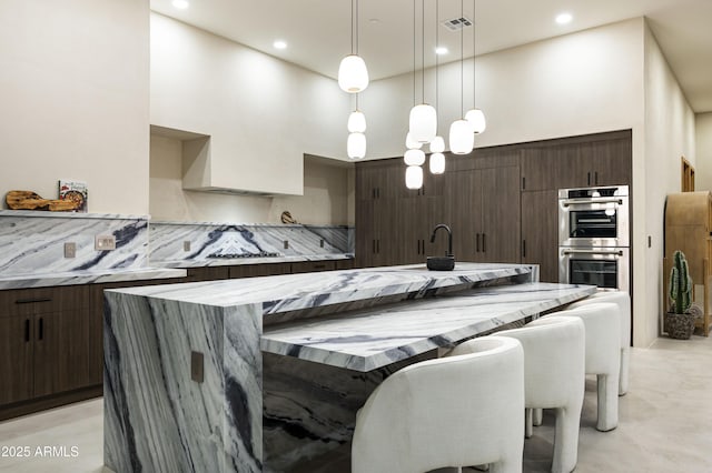 kitchen featuring decorative backsplash, a kitchen island with sink, a high ceiling, dark brown cabinets, and double oven