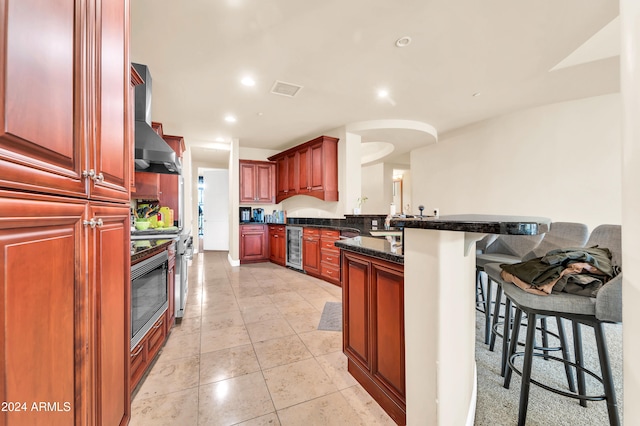 kitchen featuring wall chimney exhaust hood, kitchen peninsula, dark stone countertops, a kitchen bar, and beverage cooler