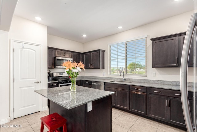 kitchen with appliances with stainless steel finishes, a sink, light tile patterned floors, dark brown cabinets, and a kitchen island