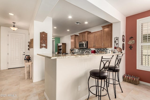 kitchen with visible vents, brown cabinetry, stainless steel microwave, a kitchen breakfast bar, and light stone counters