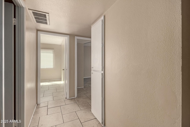 corridor featuring a textured ceiling, light tile patterned floors, baseboards, visible vents, and a textured wall
