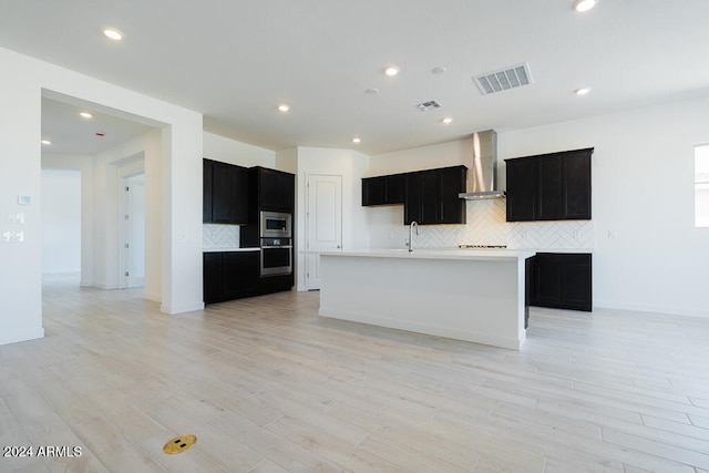 kitchen featuring a center island with sink, wall chimney range hood, stainless steel appliances, and light wood-type flooring