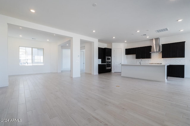 kitchen with wall chimney range hood, appliances with stainless steel finishes, an island with sink, light hardwood / wood-style floors, and decorative backsplash