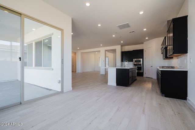 kitchen with backsplash, stainless steel microwave, a kitchen island with sink, light hardwood / wood-style floors, and sink