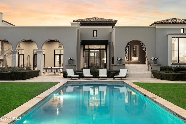 back of house at dusk with a tile roof, a lawn, stucco siding, an outdoor pool, and a patio area