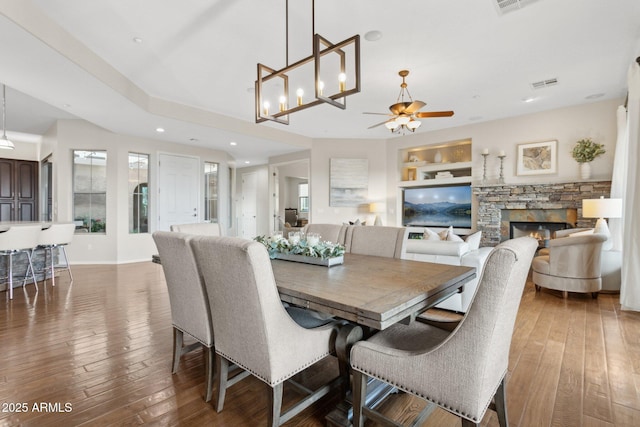 dining space featuring dark hardwood / wood-style flooring, built in shelves, a stone fireplace, and ceiling fan