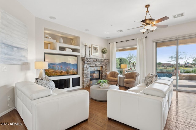 living room featuring a stone fireplace, built in features, dark hardwood / wood-style floors, and ceiling fan