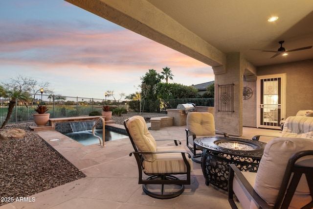 patio terrace at dusk with a fire pit, ceiling fan, area for grilling, pool water feature, and an outdoor kitchen