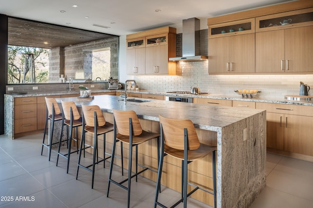 kitchen featuring sink, a kitchen island with sink, a kitchen breakfast bar, decorative backsplash, and wall chimney exhaust hood