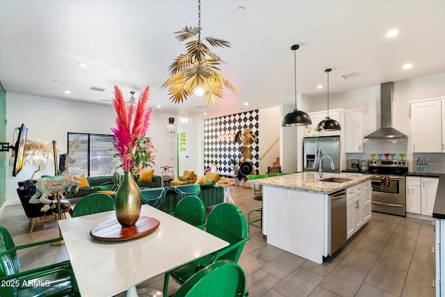 kitchen with appliances with stainless steel finishes, wall chimney exhaust hood, decorative light fixtures, sink, and a kitchen island with sink