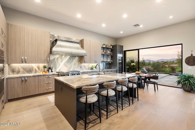 kitchen with an island with sink, sink, custom range hood, a breakfast bar area, and light stone countertops