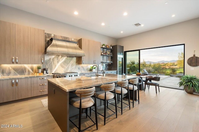 kitchen with custom exhaust hood, an island with sink, light hardwood / wood-style flooring, stainless steel range, and light stone countertops