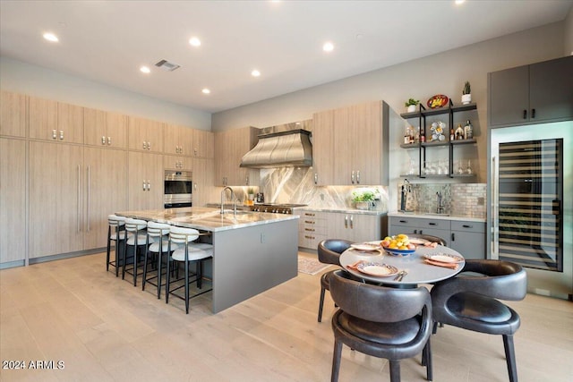 kitchen featuring a kitchen island with sink, wine cooler, light stone counters, appliances with stainless steel finishes, and premium range hood