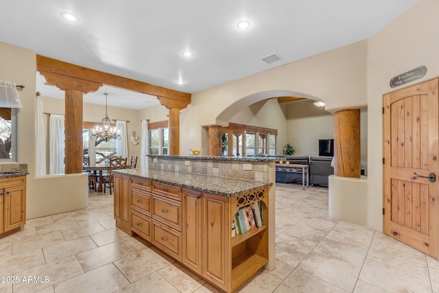 kitchen with a kitchen island, hanging light fixtures, dark stone counters, a chandelier, and ornate columns