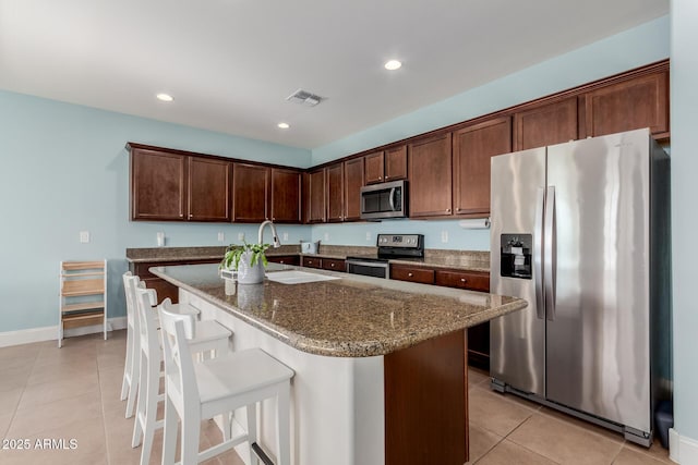 kitchen with a breakfast bar area, appliances with stainless steel finishes, dark stone counters, and a kitchen island with sink