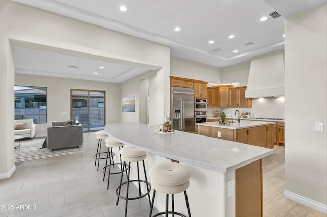 kitchen featuring an island with sink, custom range hood, appliances with stainless steel finishes, and a kitchen breakfast bar