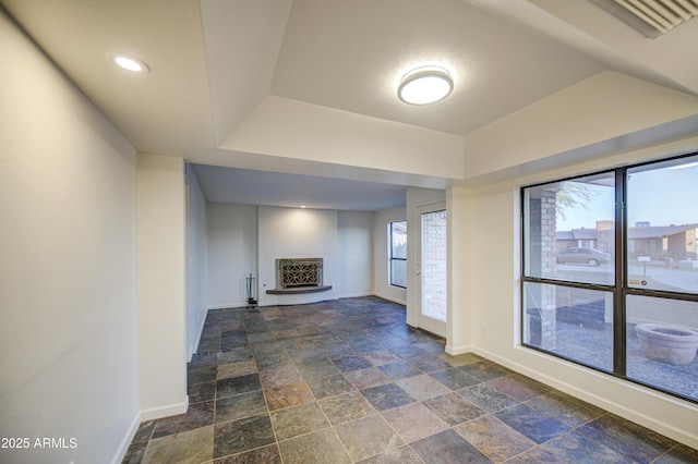 unfurnished living room with stone tile flooring, a fireplace with raised hearth, a tray ceiling, and baseboards