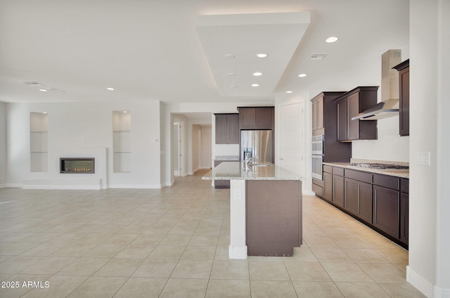 kitchen featuring an island with sink, light stone counters, stainless steel appliances, wall chimney range hood, and built in shelves