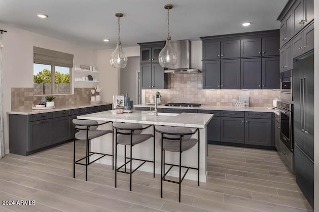 kitchen featuring stainless steel appliances, wall chimney range hood, decorative light fixtures, sink, and a kitchen island with sink