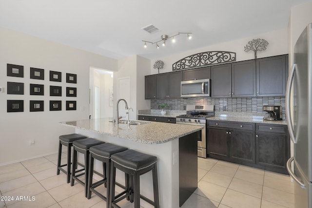 kitchen with sink, tasteful backsplash, light stone counters, an island with sink, and appliances with stainless steel finishes