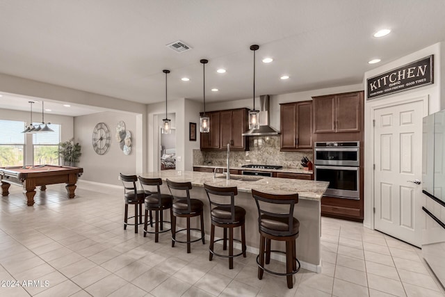 kitchen with appliances with stainless steel finishes, dark brown cabinets, pool table, wall chimney range hood, and an island with sink