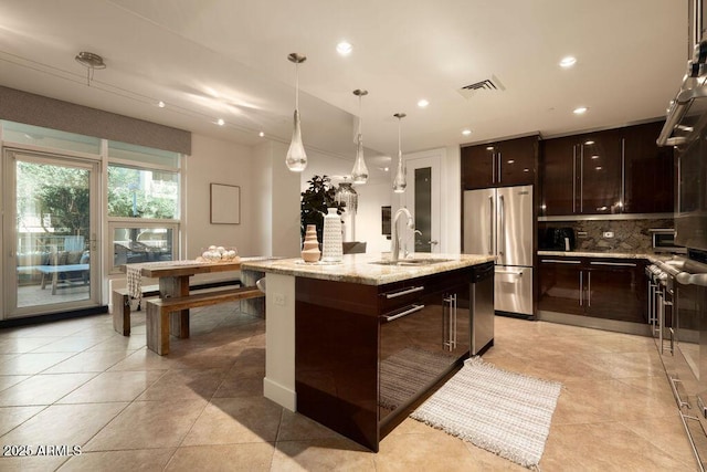 kitchen with decorative light fixtures, sink, stainless steel fridge, a kitchen island with sink, and dark brown cabinets