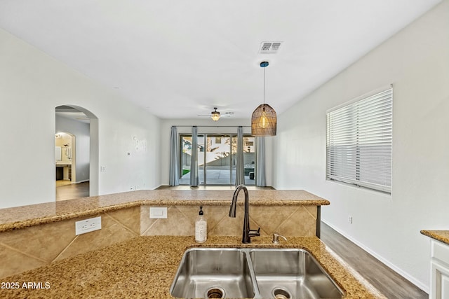 kitchen with arched walkways, light stone counters, wood finished floors, hanging light fixtures, and a sink