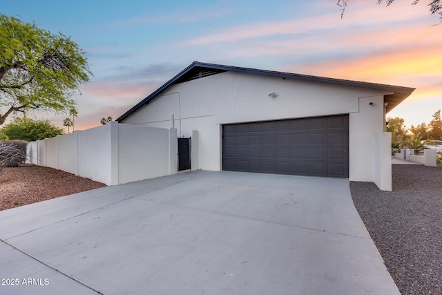 property exterior at dusk featuring fence, an attached garage, stucco siding, and concrete driveway
