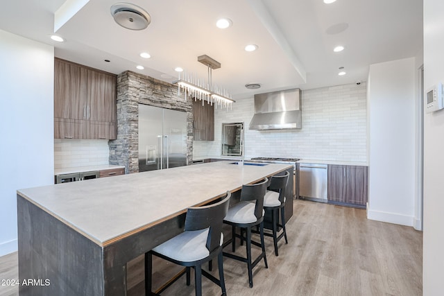 kitchen featuring wall chimney range hood, decorative backsplash, appliances with stainless steel finishes, light hardwood / wood-style flooring, and a center island