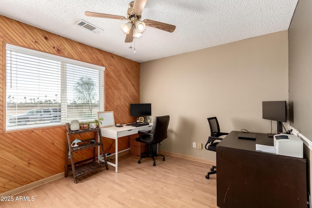 office area featuring light wood-type flooring, visible vents, a textured ceiling, wooden walls, and ceiling fan