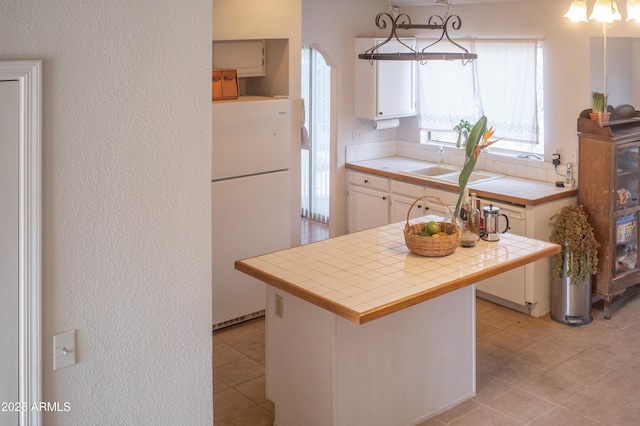 kitchen with tile countertops, freestanding refrigerator, a sink, white cabinets, and a textured wall