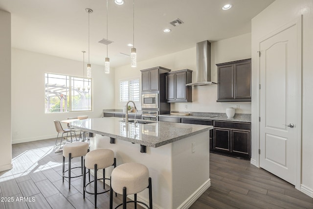 kitchen with wall chimney range hood, a center island with sink, a sink, stainless steel appliances, and a kitchen breakfast bar