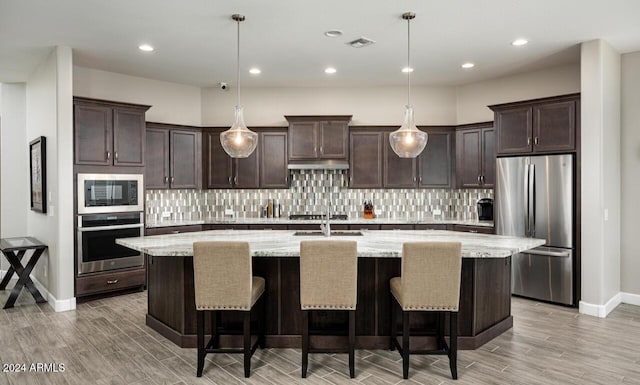 kitchen with decorative light fixtures, light stone countertops, stainless steel appliances, and a kitchen island with sink