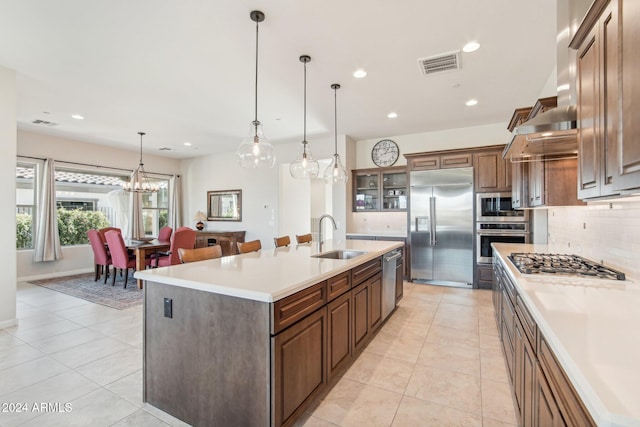 kitchen with backsplash, sink, built in appliances, decorative light fixtures, and a center island with sink