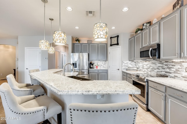 kitchen with gray cabinetry, an island with sink, a breakfast bar, appliances with stainless steel finishes, and decorative light fixtures