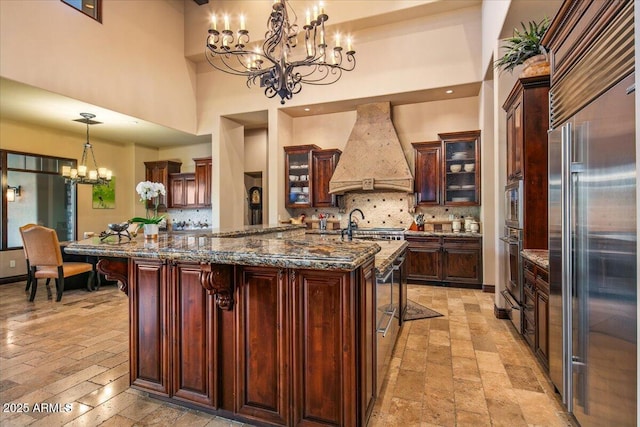 kitchen with custom range hood, glass insert cabinets, an inviting chandelier, appliances with stainless steel finishes, and decorative backsplash