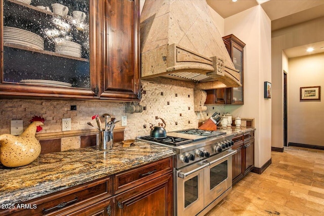 kitchen featuring dark stone countertops, double oven range, custom exhaust hood, decorative backsplash, and glass insert cabinets