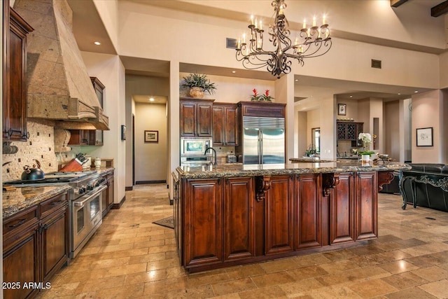 kitchen with a large island, dark stone countertops, tasteful backsplash, built in appliances, and custom exhaust hood