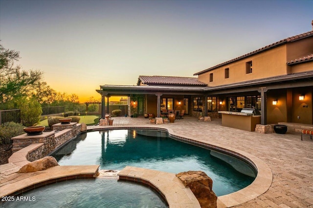 pool at dusk featuring outdoor dry bar, a pool with connected hot tub, fence, an outdoor kitchen, and a patio