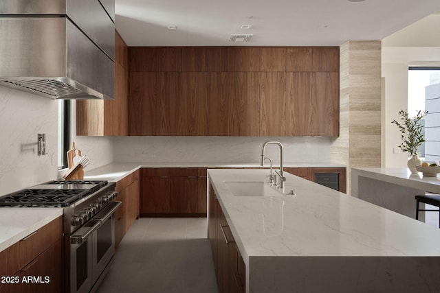 kitchen featuring a center island with sink, light stone countertops, range with two ovens, wall chimney exhaust hood, and sink