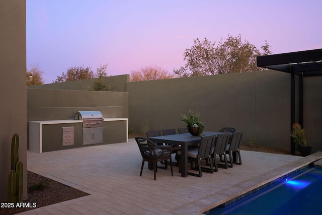 patio terrace at dusk featuring an outdoor kitchen, grilling area, and a fenced in pool