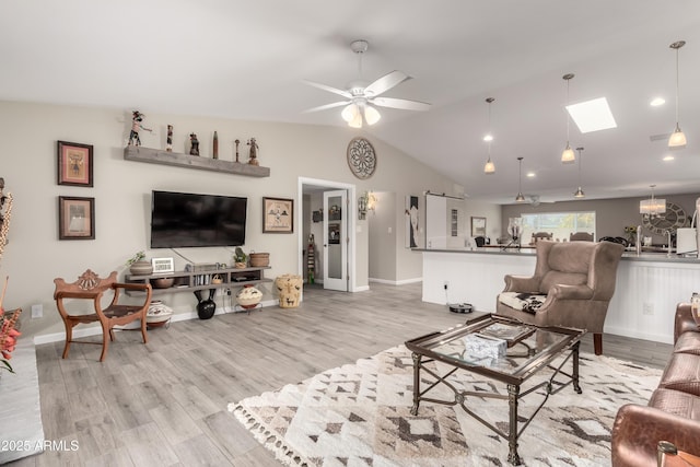 living room featuring ceiling fan with notable chandelier, light wood finished floors, lofted ceiling with skylight, and baseboards