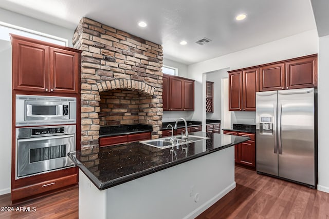 kitchen featuring an island with sink, appliances with stainless steel finishes, dark stone counters, dark wood-type flooring, and sink